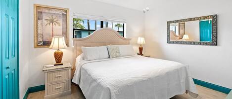 Un dormitorio con cama, dos mesitas de noche con lámparas, un ventilador de techo, una ventana con vista a palmeras y una puerta con marco color turquesa.
