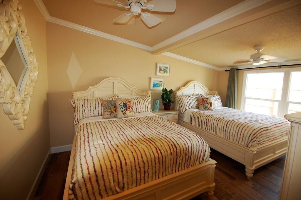 A bedroom with two beds, a ceiling fan, a large mirror, and a window with curtains.