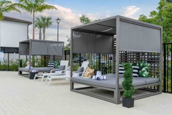 A modern outdoor seating area with cushioned chairs and a small table, surrounded by a metal fence and palm trees.