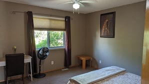 A bedroom with a bed, a desk, a chair, a standing fan, a window with curtains, a small stool, and a painting on the wall. - 6 beds entire house fenced Yard,  2 car Pad + Garage (3 beds at basement) (Calgary)