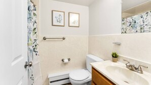 Ein Badezimmer mit Toilette, Waschbecken und Duschvorhang. An der Wand hängen zwei Bilder und auf dem Waschbecken steht eine kleine Pflanze.