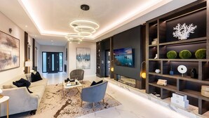 Un salon moderne avec un canapé blanc, un fauteuil gris, une table basse et un téléviseur fixé au mur. On y trouve une bibliothèque avec des objets décoratifs et un grand tableau au-dessus.