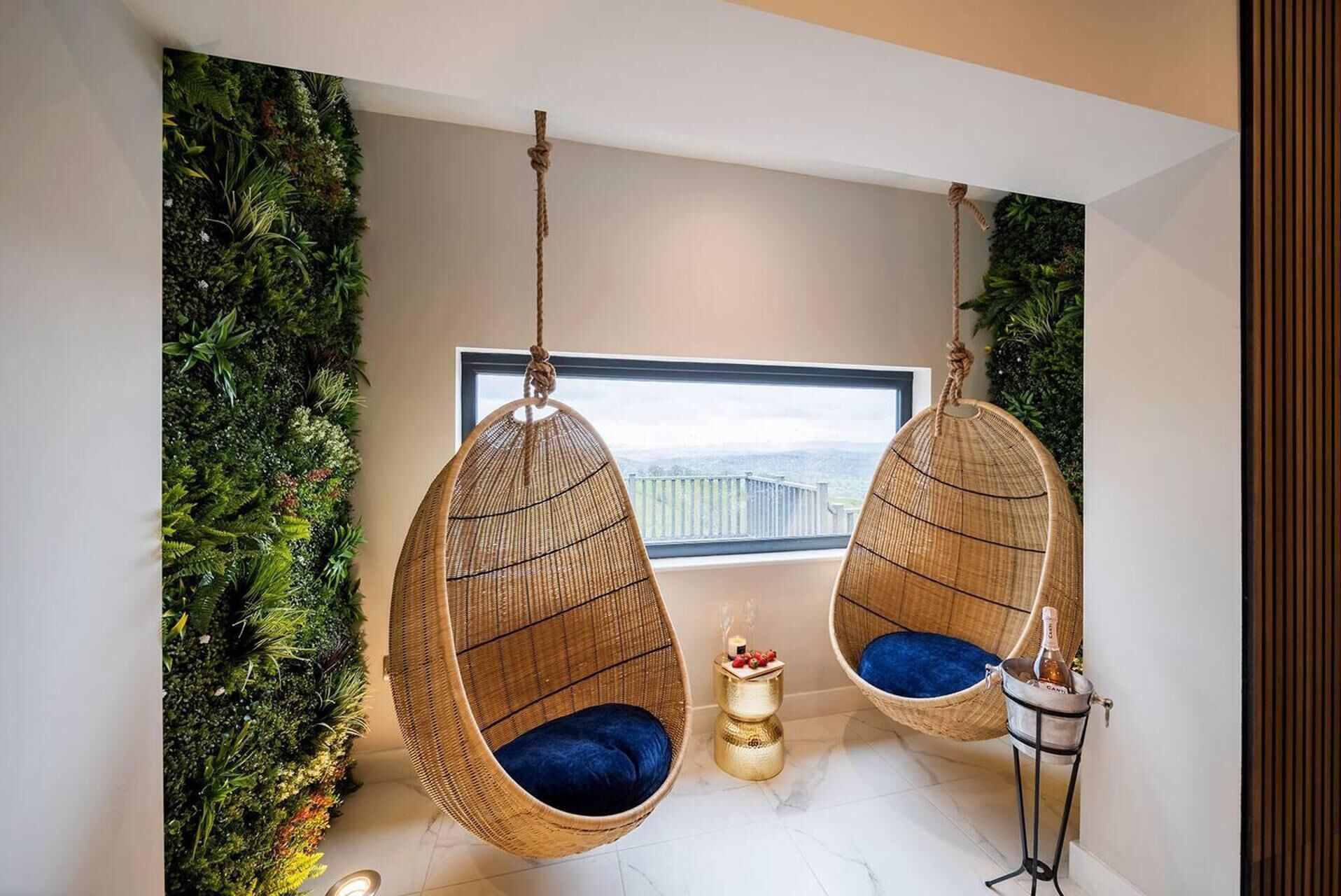 Una stanza con due poltrone in vimini appese, cuscini azzurri, un tavolino laterale dorato e vista su un balcone con vegetazione.