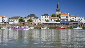Une ville côtière avec un clocher d’église imposant, des bateaux amarrés le long de la rive et divers bâtiments, dont un hôtel dont l’enseigne est bien visible.