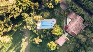 An aerial view of a property with a swimming pool, a small building with a red roof, and surrounding greenery.