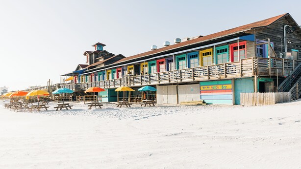 Un edificio de madera frente a la playa con toldos de colores y un letrero de bar de playa.