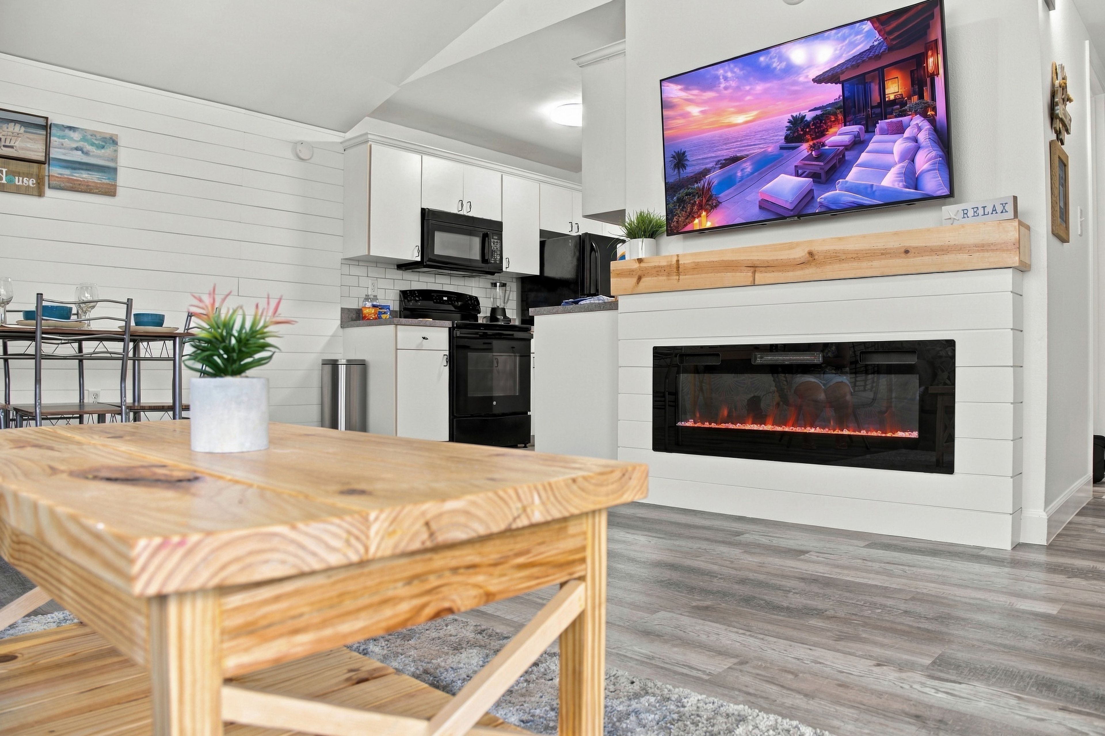 A modern kitchen with white cabinetry, a flat-screen TV mounted above the fireplace, and a dining area with a wooden table and chairs.