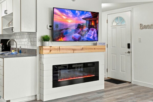 A modern kitchen with a flat-screen TV mounted above a fireplace, a white door with a window, and a kitchen counter with a sink and cabinets.