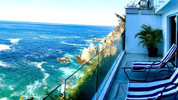 A balcony with a view of the ocean, a potted plant, and striped lounge chairs. - El acantilado (Acapulco)