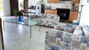 A modern living room with a glass coffee table, patterned sofas, a kitchen with wooden cabinets, and a TV. - El acantilado (Acapulco)