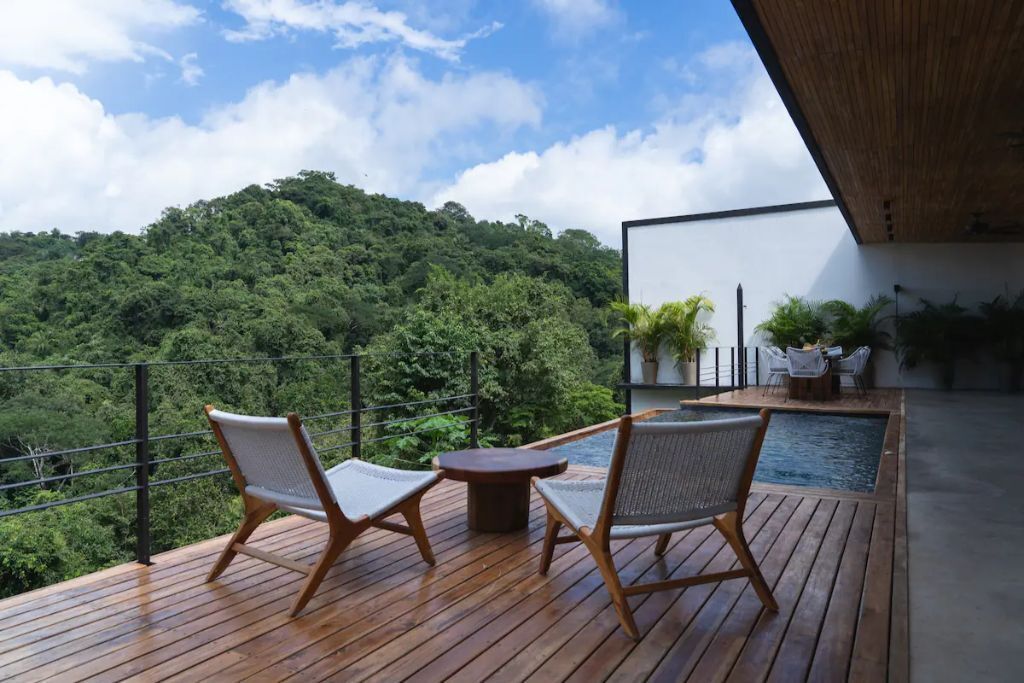 A wooden deck with two wicker chairs and a small table, overlooking a lush green forest.