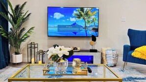 A modern living room with a large flat-screen TV mounted on the wall, a glass coffee table with decorative items, a blue armchair, and a potted plant. - Newly updated Modern 2 BR  Scottsdale Condo. (Scottsdale)