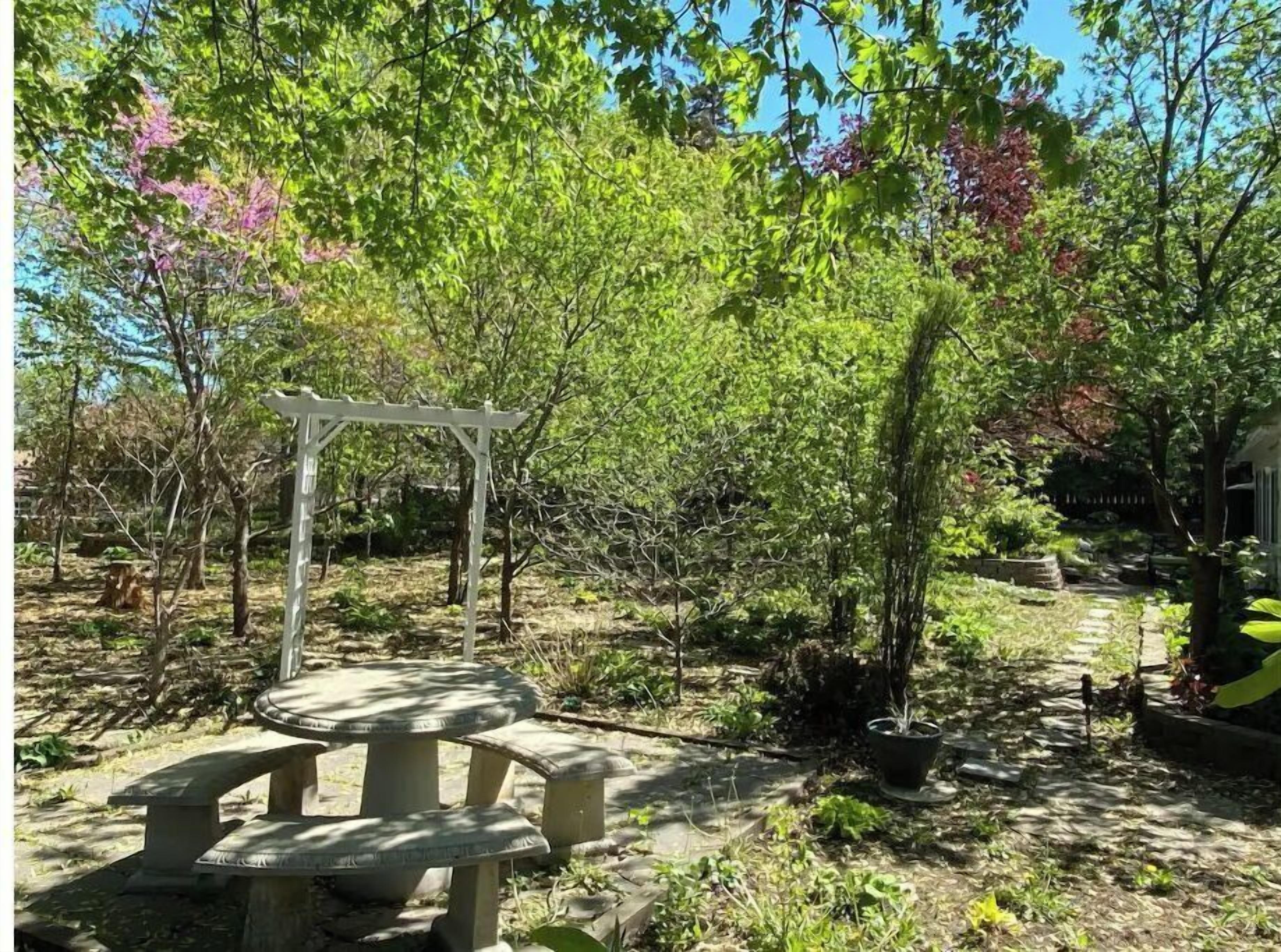 Ein Garten mit einer weißen Pergola, einem runden Steintisch und unterschiedlichen Pflanzen.