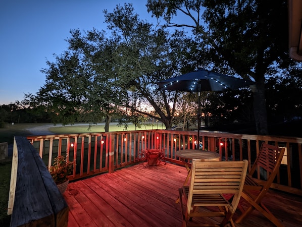 Une terrasse en bois avec une table et des chaises, entourée d’une rambarde et illuminée de guirlandes lumineuses.