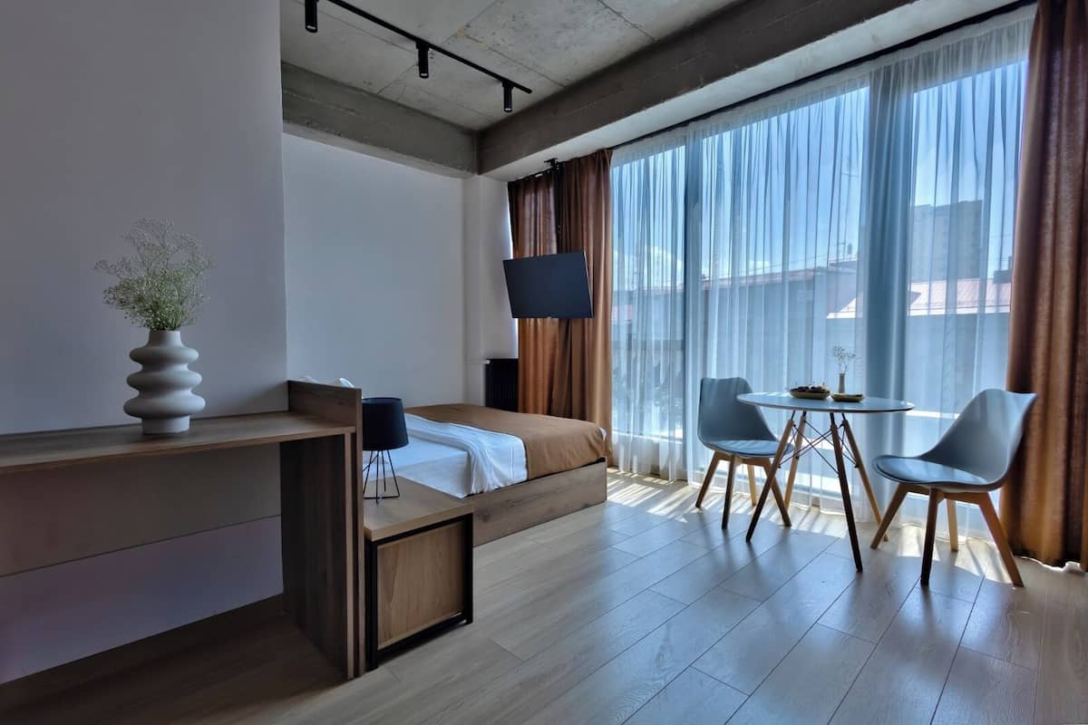 Quarto de hotel com cama, escrivaninha, cadeira, televisão e janelas amplas.