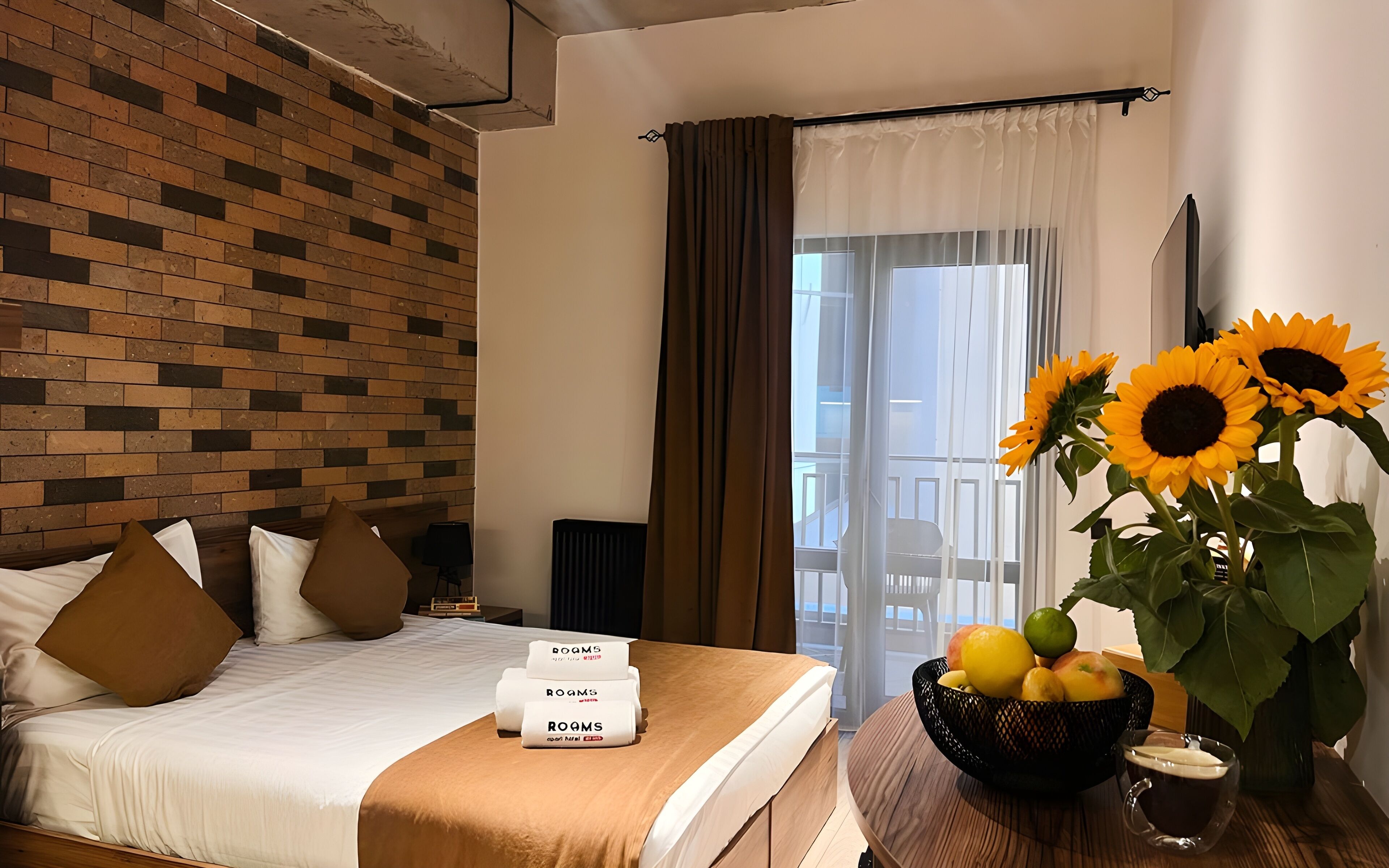 A hotel room with a brick-patterned wall, a bed with brown pillows, a bedside table with a bowl of fruit and a glass of drink, and a window with curtains.