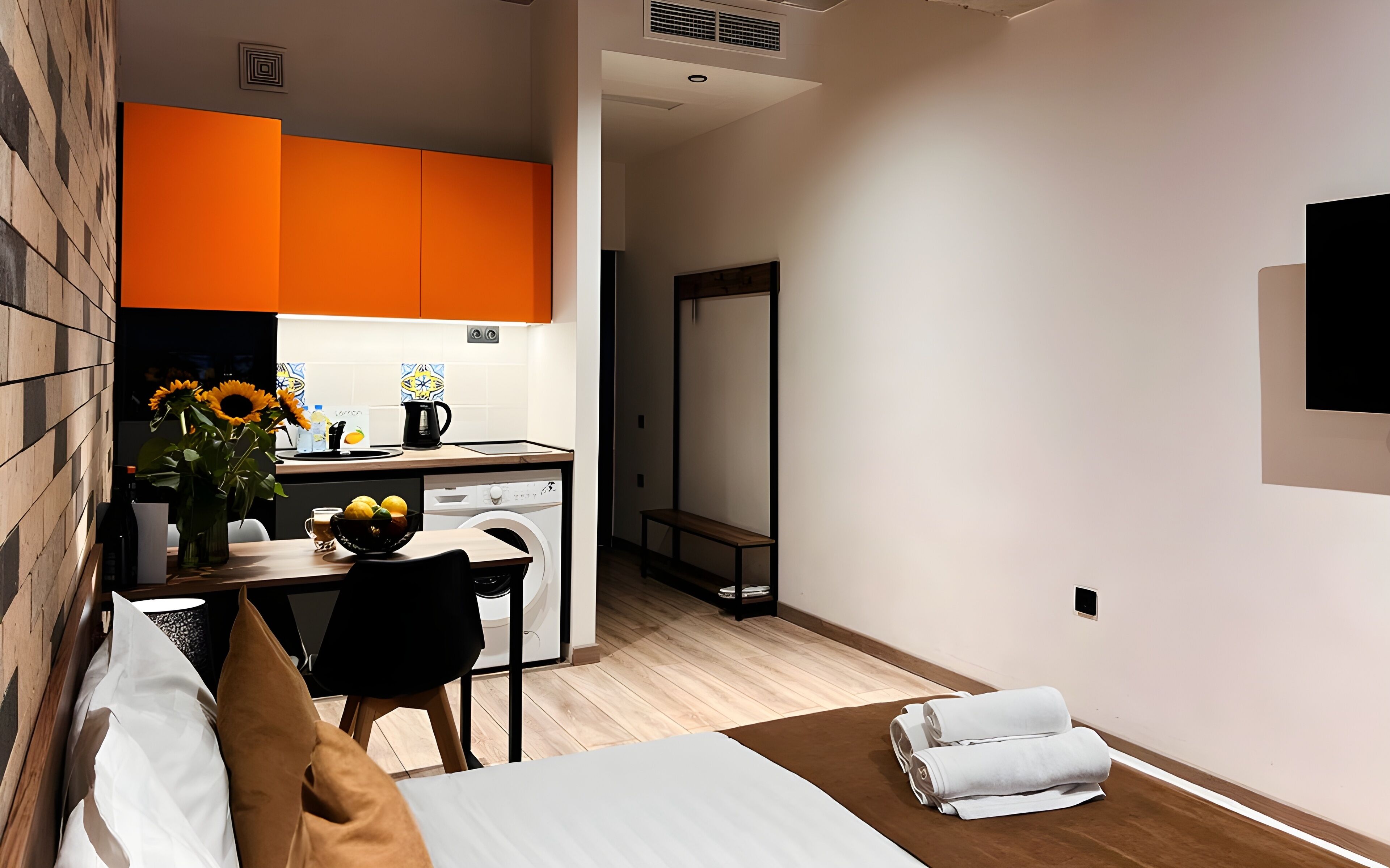 A modern hotel room with a bed, a small desk with a chair, a dining table with a vase of sunflowers, and a washing machine.