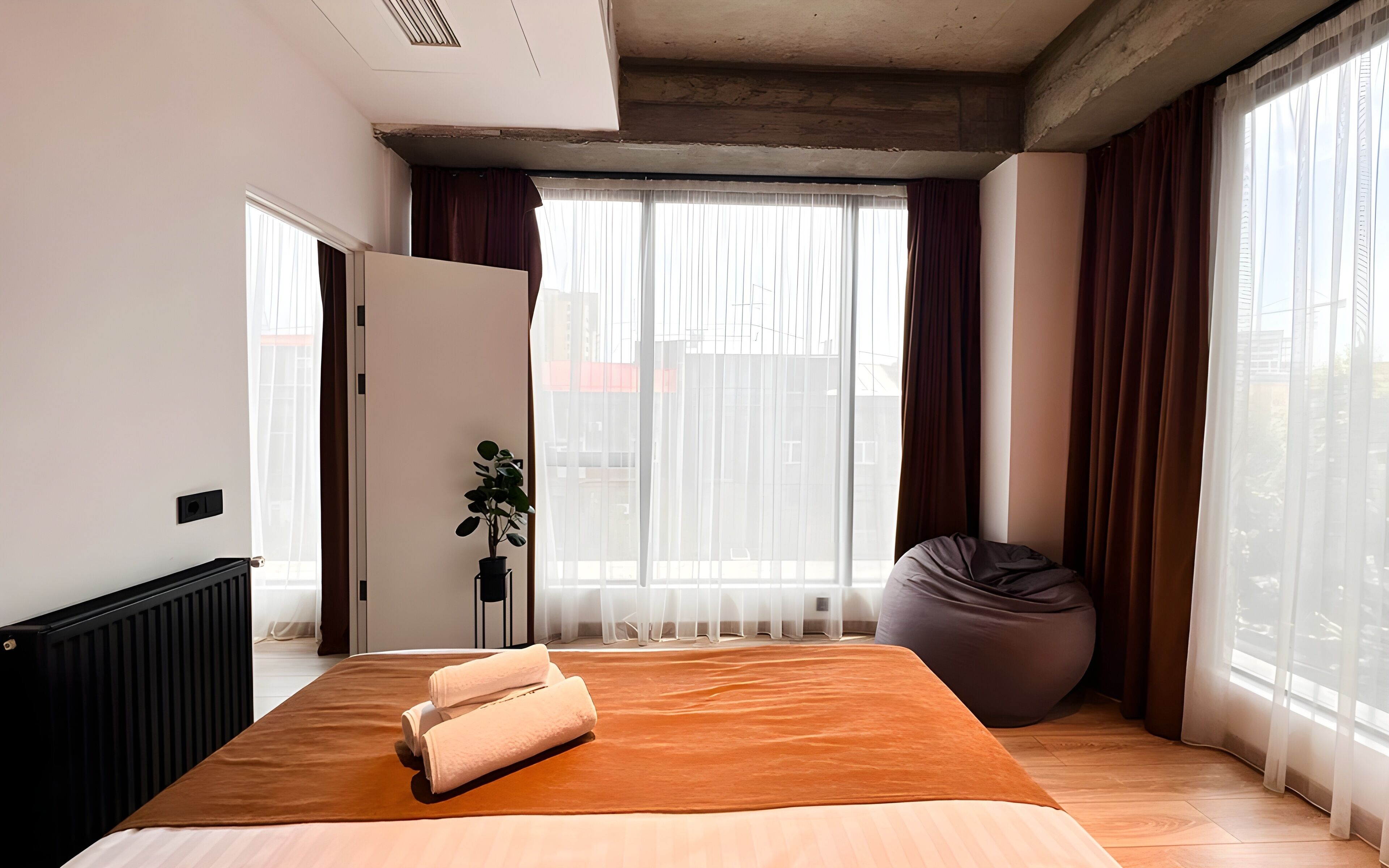 A modern bedroom with a large bed, a potted plant, a bean bag, and large windows with curtains.