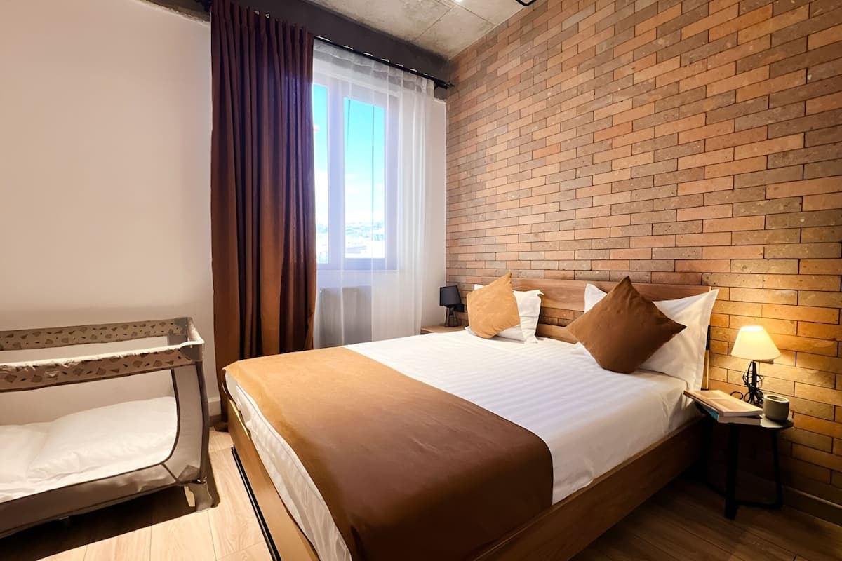 Quarto de hotel com parede de tijolos, uma cama grande com cabeceira de madeira, uma mesa de cabeceira com abajur e uma janela com cortinas.