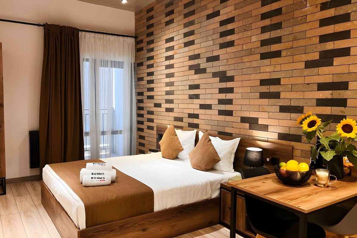 Quarto de hotel moderno com parede em padrão tijolo, cama de madeira com roupa de cama branca e escrivaninha de madeira com uma tigela de frutas e uma luminária.