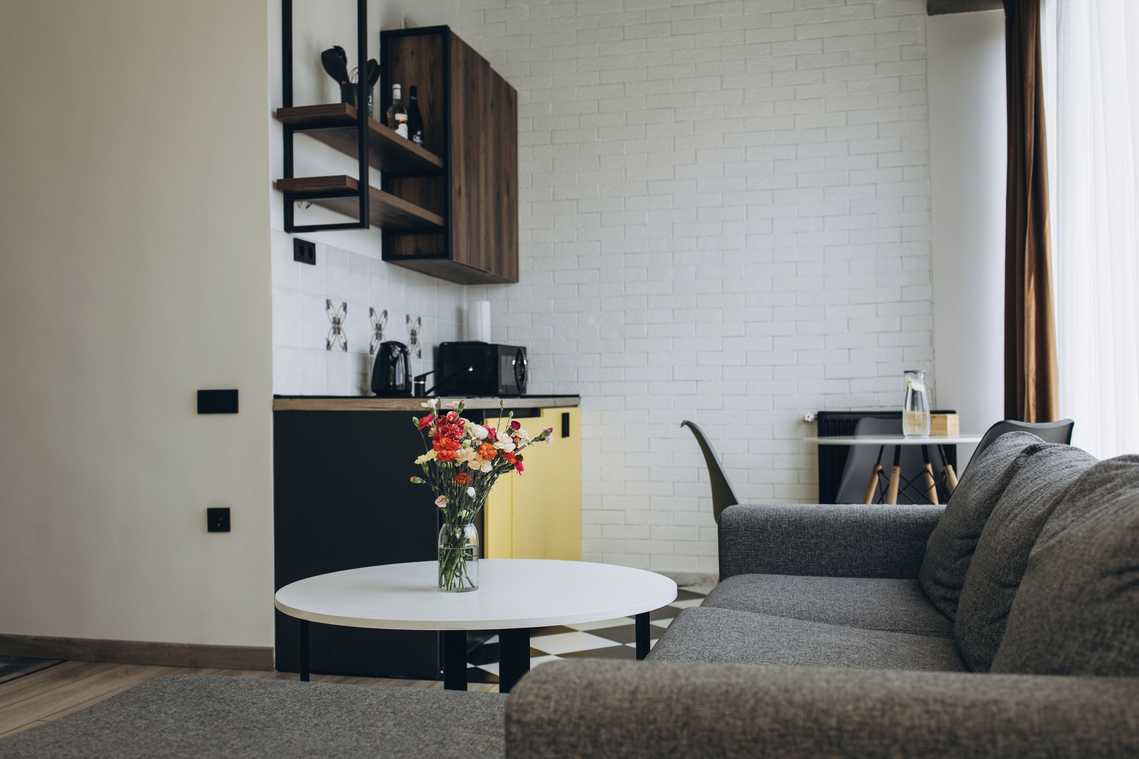 A modern living room with a sofa, a coffee table with a vase of flowers, a kitchen area with a yellow cabinet, and a brick wall.
