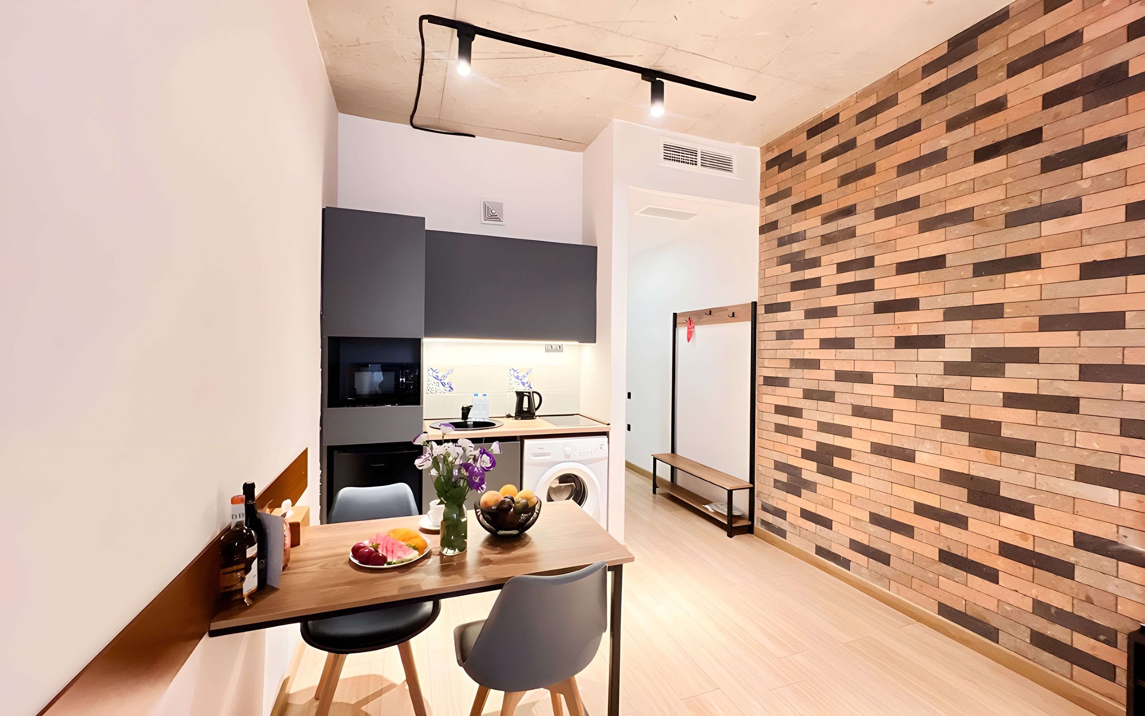 A modern kitchen with a built-in oven, a microwave, and a coffee machine. There is a dining area with a table and chairs, and a brick-patterned wall.