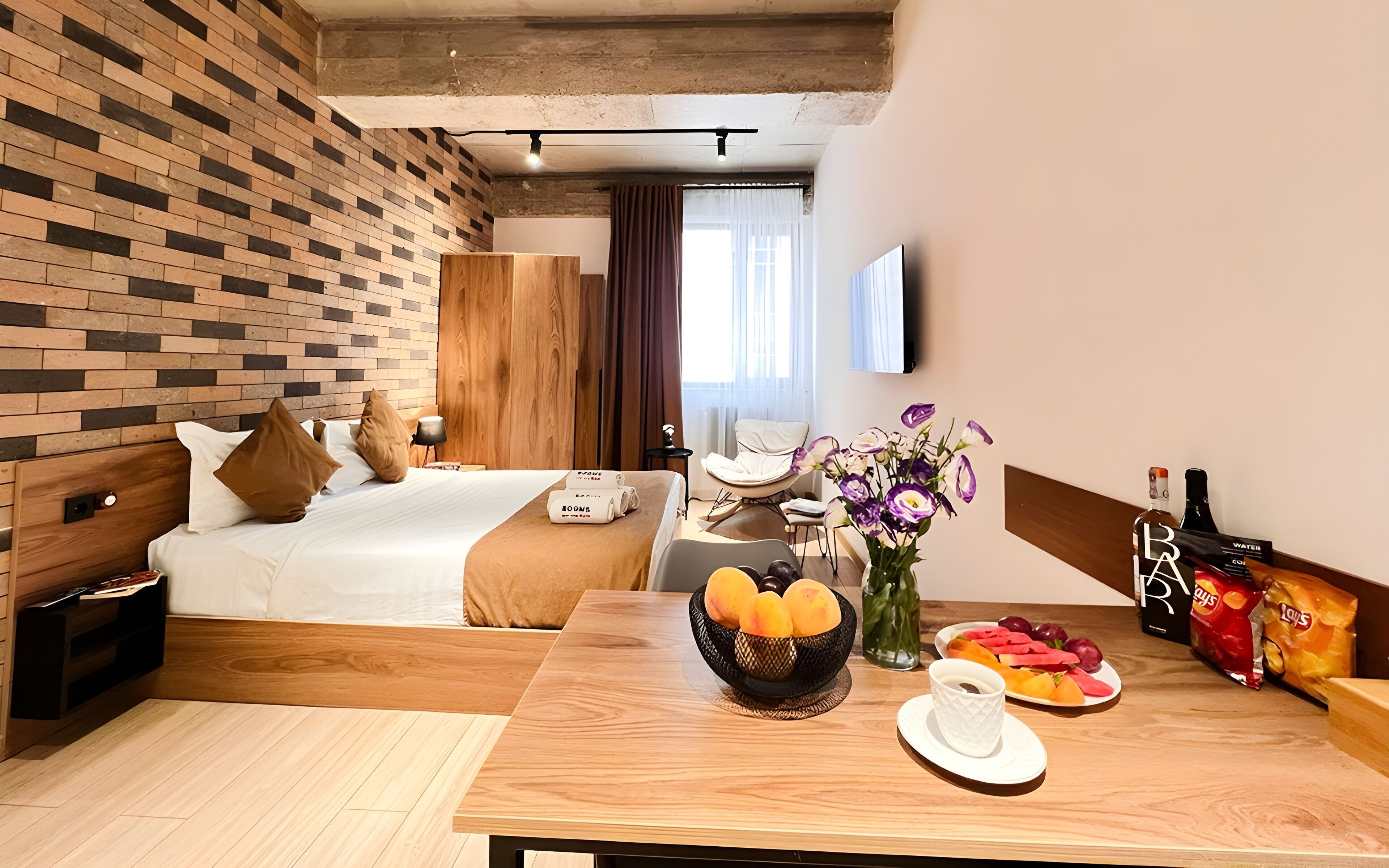 Une chambre d’hôtel avec un lit, un bureau, une table à manger avec des fruits et des en-cas, un vase avec des fleurs et une bouteille d’eau.