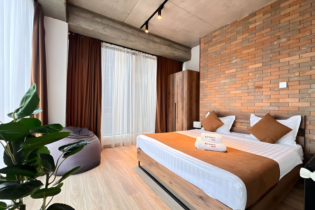 Quarto de hotel com parede de tijolos, cama com roupa de cama branca e almofadas marrons, piso de madeira e uma janela grande com cortinas.