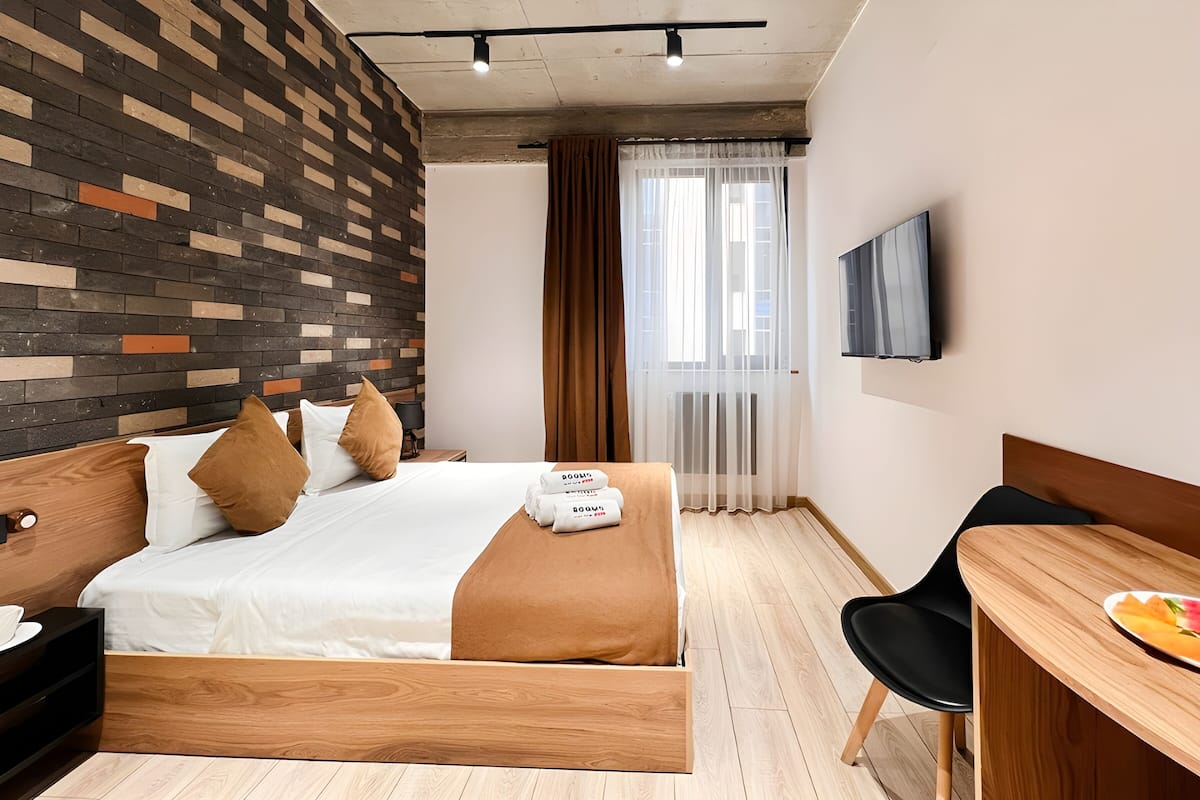 Quarto de hotel com cama, escrivaninha, cadeira, televisão e janela com cortinas.