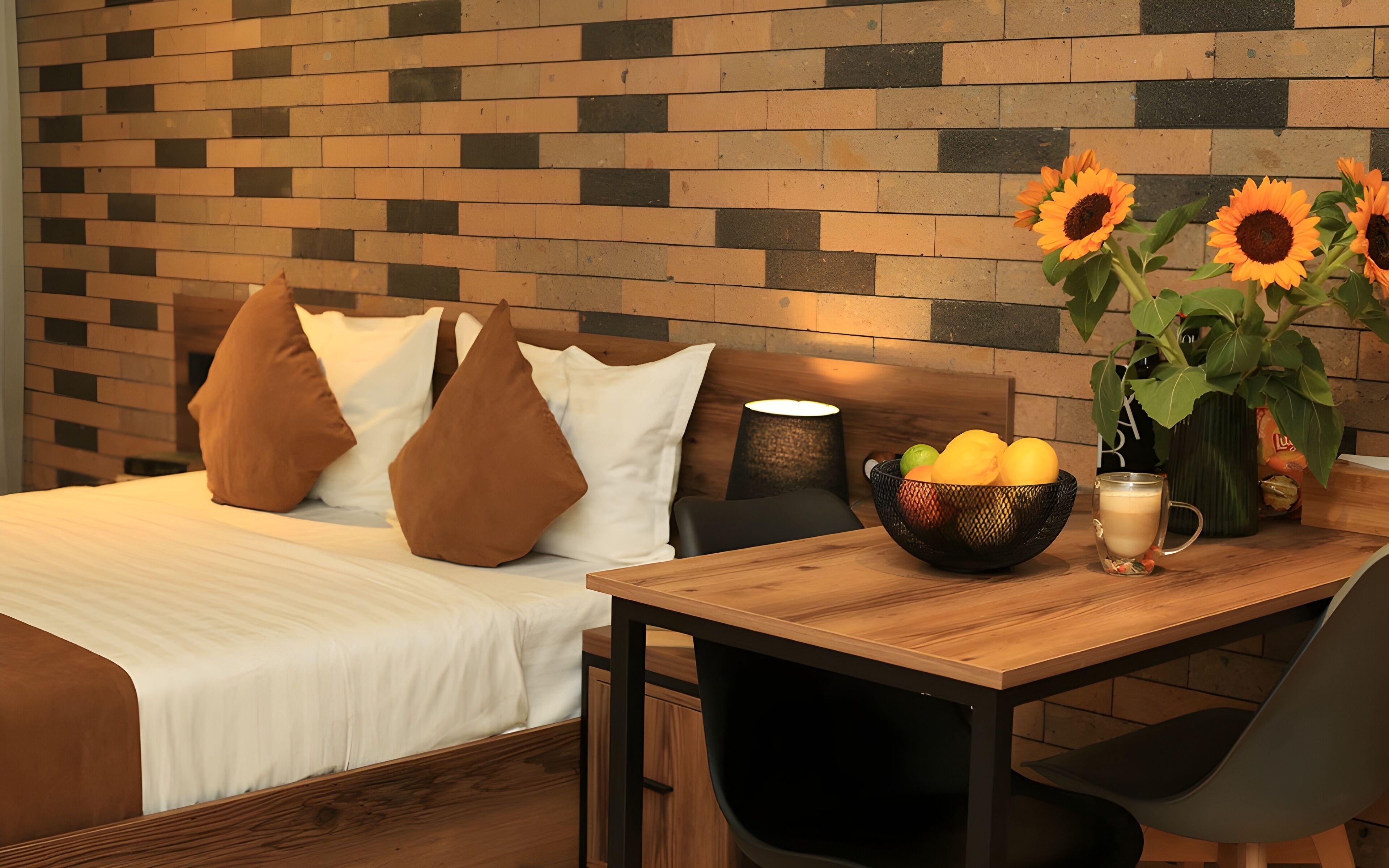 A neatly arranged hotel room with a bed, a wooden table with fruits and a vase of sunflowers, and a brick-patterned wall.