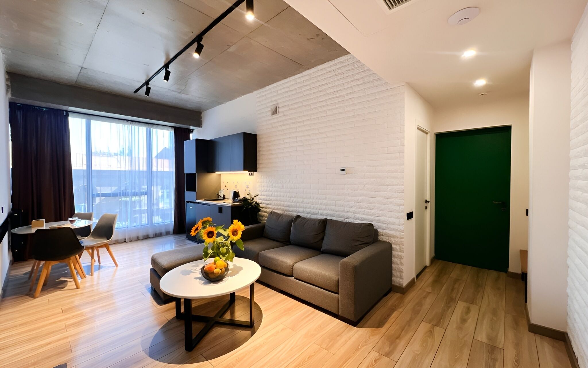 A modern living room with a sofa, coffee table, and a dining area with chairs. There is a kitchen area with a counter and a green door.