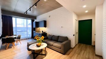 A modern living room with a sofa, coffee table, and a dining area with chairs. There is a kitchen area with a counter and a green door.