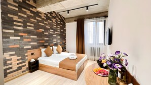 Une chambre d’hôtel moderne avec un lit en bois, un mur décoré d’un motif en briques, une table de chevet en bois, une petite table à côté du lit ornée d’un vase de fleurs et une assiette de fruits.