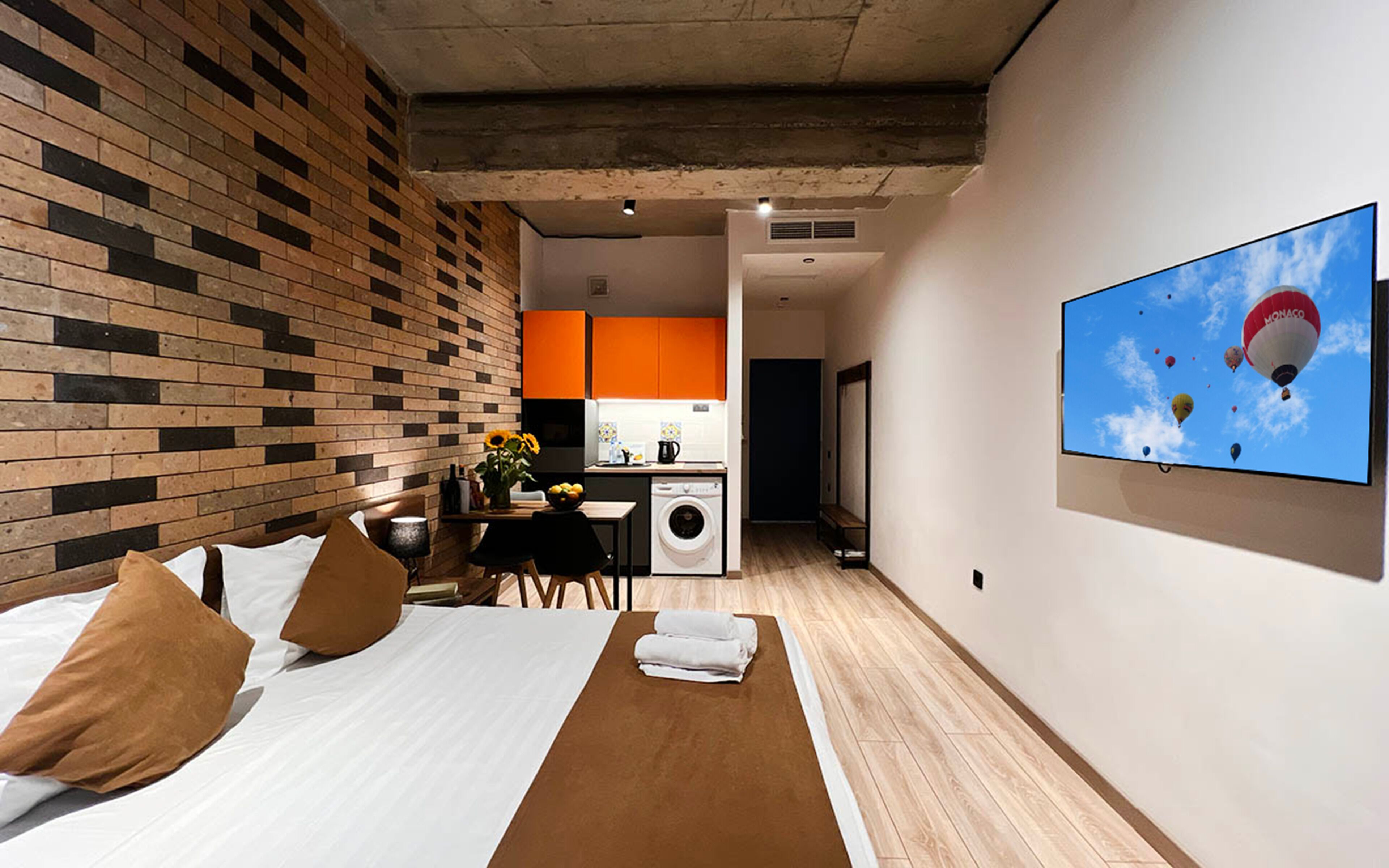 A hotel room with a brick-patterned wall, a bed with white linens, a wooden bedside table, a small table with a vase of sunflowers, a washing machine, and a large TV screen displaying hot air balloons.