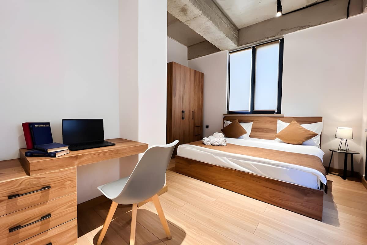 Quarto moderno com escrivaninha de madeira, um laptop e livros, uma cama com almofadas e uma mesa de cabeceira com abajur.