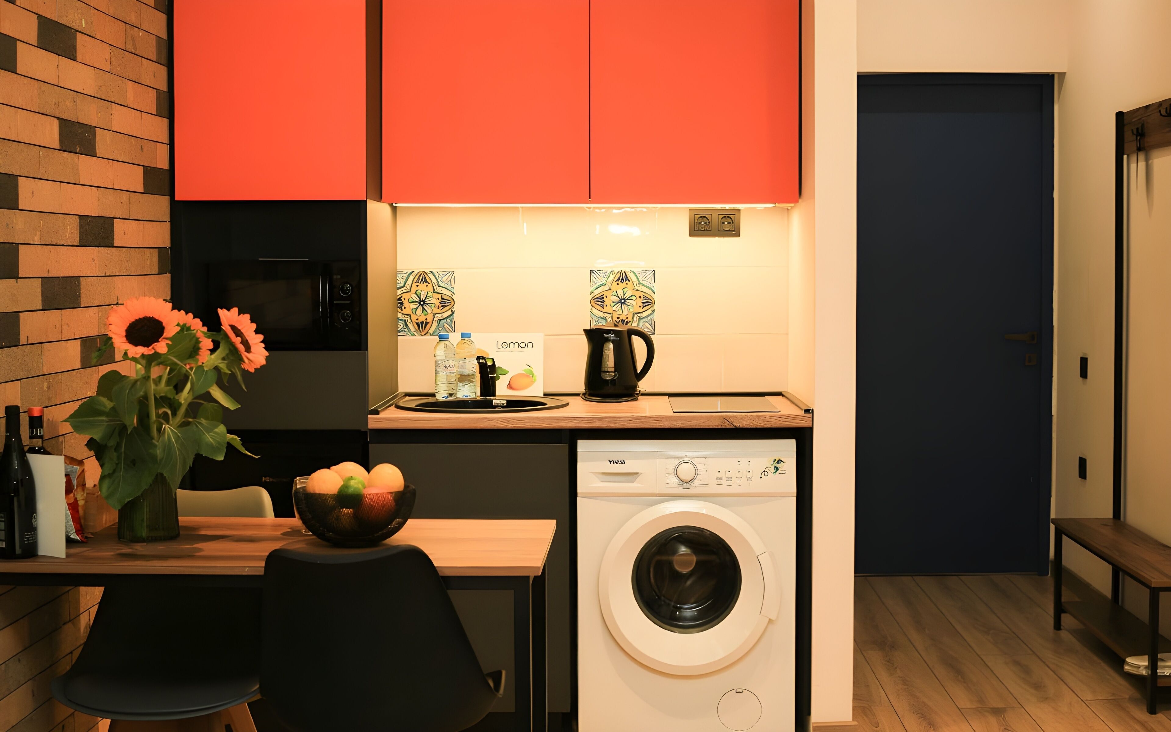 A compact kitchen with a washing machine, a dining table with chairs, and a vase with flowers.