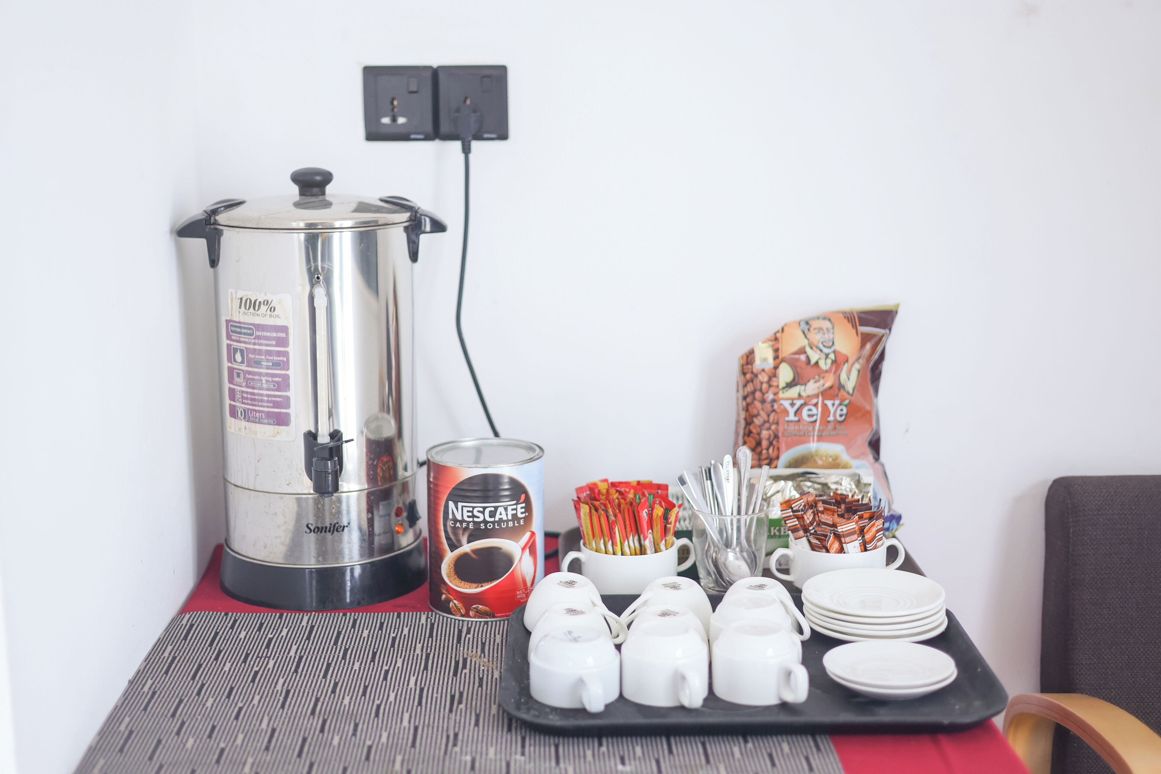 Una estación de café con cafetera Nescafe, vasos desechables, teteras y una bandeja con platos y cubiertos.