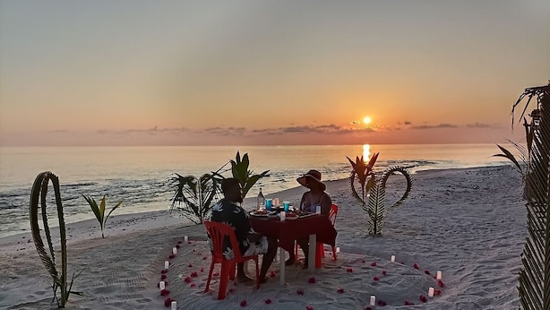 Une scène de plage au coucher du soleil, avec une table dressée pour deux personnes, entourée de bougies et de feuilles de palmier.