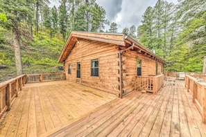 A wooden cabin with a deck, surrounded by forest.