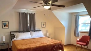 Een slaapkamer met een bed, een plafondventilator, een stoel, een raam en een schilderij aan de muur.