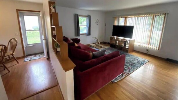Een ruime zithoek met een rode bank, houten vloeren, een flatscreen televisie en een groot raam met zonwering.