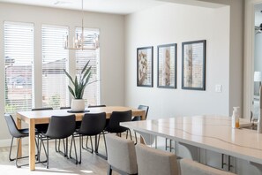 Un comedor moderno con una mesa de madera y sillas negras, ventanas amplias con persianas y tres obras de arte enmarcadas en la pared.