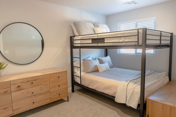 Une chambre avec un lit superposé, une commode en bois, un miroir rond et une fenêtre avec des stores.
