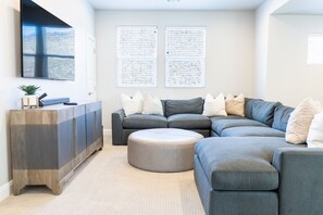 A modern living room with a sectional sofa, a round ottoman, a wooden cabinet, and two large windows with artwork.