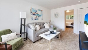 Ein modernes Wohnzimmer mit einem grauen Sofa, einem weißen Couchtisch und einem grünen Sessel. Ein hoher, schwarzer Bücherregal und ein Gemälde sind an der Wand zu sehen.