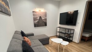 A modern living room with a grey sectional sofa, a flat-screen TV, a small white table, and a wooden floor. - Charming Bas de villa beach walking natural site (Venzolasca)