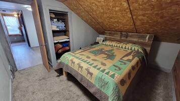 A bedroom with a sloped ceiling, a bed with a patterned quilt, a wooden headboard, and a closet with folded blankets and pillows.