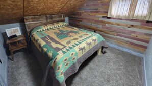 Un dormitorio con techo inclinado, paneles de madera en la pared, una cama con colcha estampada, una mesita de noche con un marco de fotos y una ventana con cortinas.
