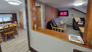 Una habitación con un espejo de marco de madera que refleja un comedor con una mesa y sillas, un sofá con una manta y una ventana con cortina roja.