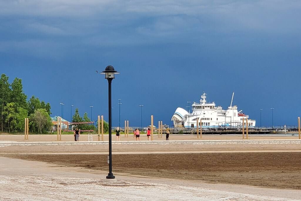 A waterfront area with a large white cruise ship docked, a promenade with people walking, and a street lamp in the foreground.